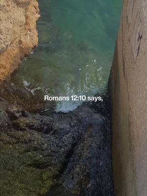 An overhead shot captures a seascape with a rocky shoreline. A stone wall forms the right edge, and a section of rocky terrain frames the left. Turquoise water fills the scene, with text "Romans 12:10 says," overlaid at the bottom.