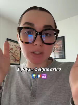 A woman wearing glasses is taking a selfie, gesturing with open hands. The background includes art pieces and a window. The text overlay says: 1 pays = 1 signe astro, accompanied by globe, French flag, and Libra symbol emojis.