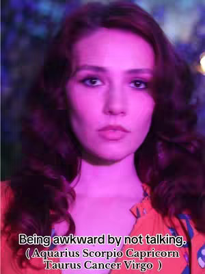 A portrait shot displays a woman with long, wavy auburn hair and a pale complexion. The lighting is bright, with a strong pink or purple hue that subtly highlights her features. The woman looks directly at the camera. At the bottom, text overlayed on the image suggests the video's focus on astrology and zodiac signs.