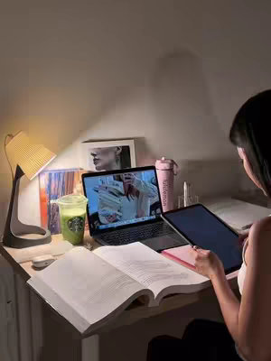 The image shows a cozy desk setup with a laptop open, revealing a video on the screen. The desk is illuminated by a light, next to an open book. A drink and a pink water bottle labeled "Lululemon" is also placed on the desk. A person is seen from the back. The overall aesthetic is warm and inviting.