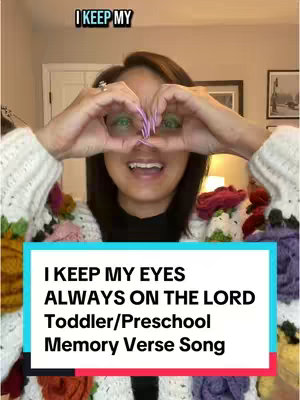 The cover image features a woman with dark hair, smiling at the camera with her hands forming a heart shape over her eyes. She has pink painted nails. Behind her, the background is blurred, featuring a neutral-toned interior with a framed picture and a hint of a lamp. Overlayed at the bottom is a light blue rectangle with a white border containing text describing the video as a toddler/preschool memory verse song.
