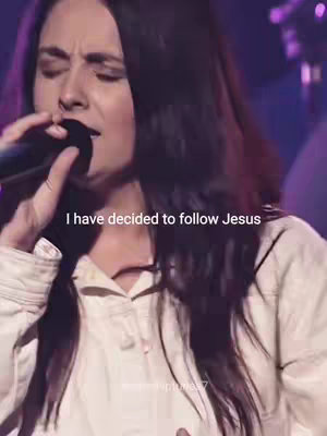 A woman with long dark hair is singing into a microphone on a stage. She wears a white top. Text overlay reads, 'I have decided to follow Jesus'. The background is blurred and dark, suggesting a concert or performance setting.
