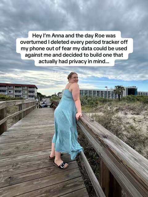 A woman in a blue dress is standing on a wooden boardwalk that extends towards the horizon. She is looking over her shoulder towards the camera with a smile. The sky is overcast, and the boardwalk leads to some buildings in the distance.