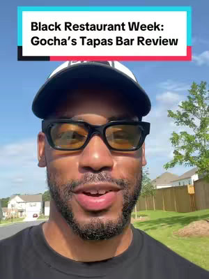 A selfie-style shot featuring a man wearing a black cap, glasses, and a black shirt, set against a bright blue sky, trees, and residential buildings. Text overlays appear at the top. The man has a beard and appears to be speaking or recording a video.