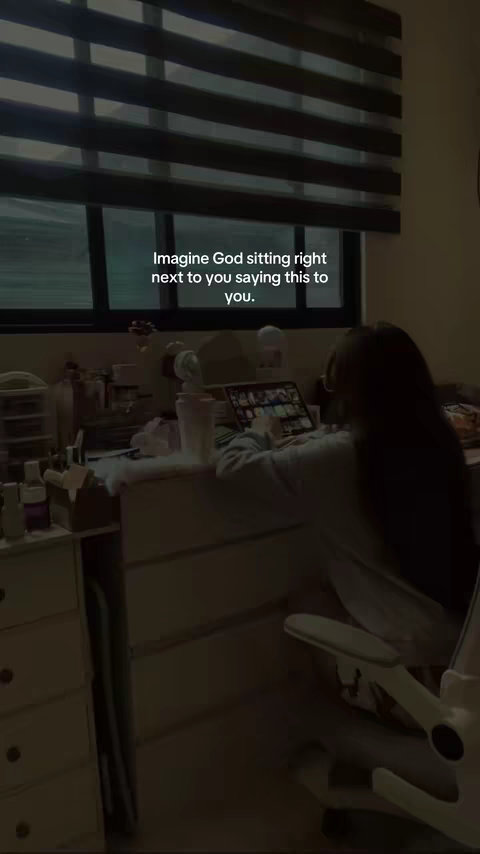 A person is working on a laptop in a dimly lit room. The person is sitting at a desk with a window behind them, partially covered by blinds. Text overlayed on the image says, 'Imagine God sitting right next to you saying this to you.'