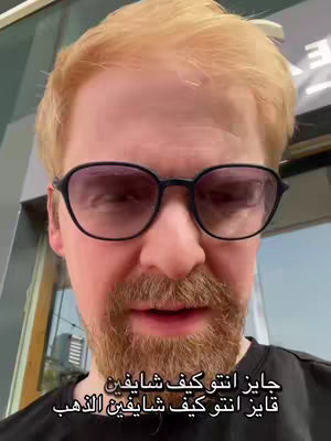 A man with light-colored hair and a reddish beard is the central figure, wearing black-rimmed glasses. The image is a close-up, focusing on the man's face from the chest up. There's text in Arabic at the bottom of the image.