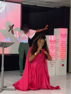 The cover image shows a stage setup with two women. A woman in a long, pink dress kneels and speaks into a microphone. She is the central focus. Behind her, another woman stands with arms outstretched, also holding a microphone. Pink columns with text are to the right of the kneeling woman, and a screen projects onto a light pink background. The overall vibe is one of celebration and religious expression.