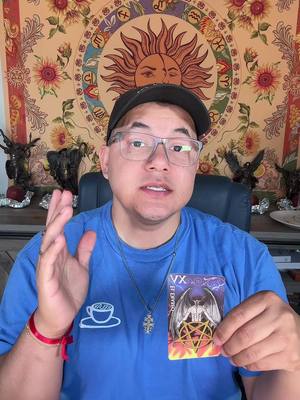 A man wearing glasses and a black cap is holding a tarot card in front of him. He's sitting in a chair, and a tapestry with a sun design hangs behind him. The man is looking at the camera, and his left hand is raised. The tarot card depicts a dark figure with wings in front of a pentagram.