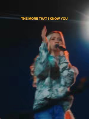A woman with blonde, shoulder-length hair is on stage, holding a microphone and singing. She's wearing a patterned jacket over a white top. The background is dark with blue and yellow lighting.