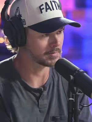 A man with a beard and a cap that says "FAITH" is speaking into a microphone while wearing headphones. The background is out of focus with a blue and purple glow.