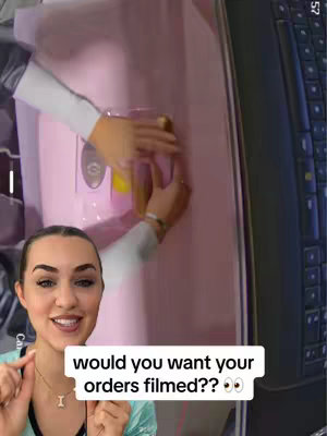 A woman is smiling directly at the camera in a selfie layout. She gestures with her fingers at a lower box that says, "Would you want your orders filmed?? 🧐🧐". In the background, a pink box is visible and there is a computer monitor and part of a keyboard on the right.