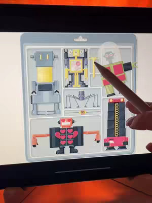 A close-up shot of a touch screen device displaying a tray with toy robot designs in various styles and colors, with a stylus pointing at one detail. The design tray is gray, and each robot is contained in its own compartment. The robots include a simple gray one, and some more elaborate ones in yellow and red. The screen is black, the stylus is dark wood with a light tip, and a human hand is barely visible.