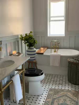 A well-lit bathroom with a bathtub, toilet, and sink. The bathroom has white walls, a white ceiling, a large window, black and white tiled flooring. There is a vase of green plants on a black toilet. The scene looks stylish and clean.