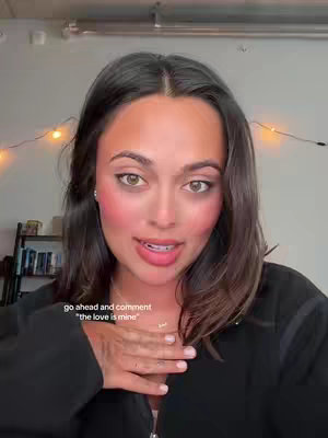 A woman with dark hair and makeup is shown from the chest up indoors, with a hand placed over her chest and text overlaid on the bottom left of the image.