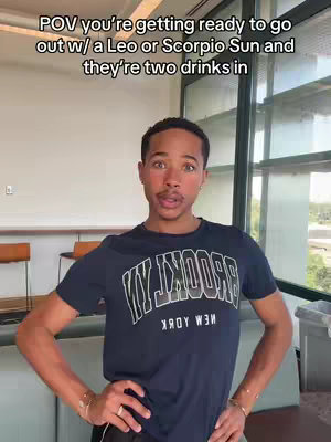 The cover image shows a young man with fair skin, dark hair and expressive eyebrows. He is positioned in front of a window with a large plant in the background. The text overlay reads "POV you're getting ready to go out w/ a Leo or Scorpio Sun and they're two drinks in". The man is wearing a dark-colored T-shirt with a "BROOKLYN" logo. The background is lit with natural light, and appears to be an office or public space. The man has his arms in a confident yet relaxed pose.