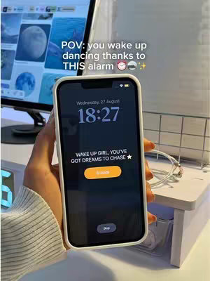 A hand holding a smartphone displaying a dark screen with white text. Above the phone is text and emojis. The screen shows the time and a message: 'WAKE UP GIRL, YOU'VE GOT DREAMS TO CHASE'. In the background is a computer monitor and a white desk setup.