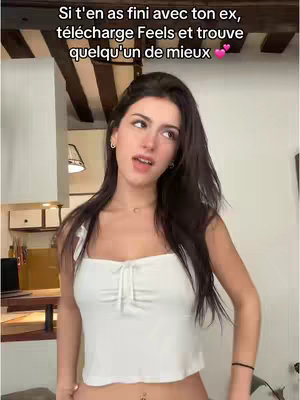 A young woman stands in a domestic setting, likely her home. She is dressed in a white top. The background consists of a kitchen area with light wood paneling and a view of a window. She is looking up and to her left with a slight smile.