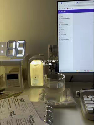 A well-lit, aesthetically composed desk setup for studying, featuring a digital clock, an illuminated decorative lamp, a glass of water, and work materials like papers and a notebook. A computer screen displaying content is also in the frame.