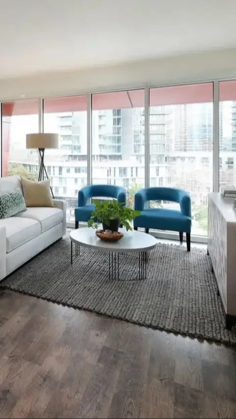 A modern living room with large windows overlooking a cityscape. The room is furnished with a white sofa, two blue armchairs, an oval coffee table, a rug, and a wooden floor. The overall impression is of a well-designed, contemporary apartment.