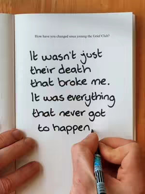 The cover image shows a person's hand holding a pen and writing on the right page of an open book. The text in black handwritten on the white paper reads, "It wasn't just their death that broke me. It was everything that never got to happen."