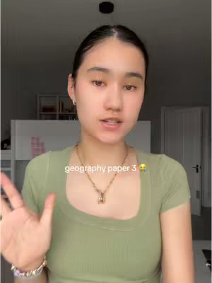 A young woman is centered in the frame with fair skin, dark hair, and a concerned expression. She is wearing a sage green scoop-neck shirt and a gold necklace. A gold chain bracelet is on her left wrist. The background features a closed door, a white cabinet, and a light fixture, suggesting an indoor setting. Text is overlayed on the bottom of the frame.