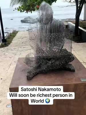 The image prominently features a metal sculpture of a seated figure, resembling a person deep in thought or meditation. The sculpture is composed of what appears to be numerous thin metal rods or wires, giving it a textured, almost pixelated appearance. The figure is positioned on a brown rectangular base. The background shows a coastal scene with water and a glimpse of a building, suggesting an outdoor setting. The text overlay contains a statement about Satoshi Nakamoto.