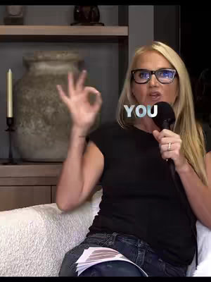 A woman with blonde hair and glasses is holding a microphone in one hand and gesturing the 'okay' symbol with the other. Overlayed text reads 'YOU'. The background includes a soft couch, shelves, and decorative items.