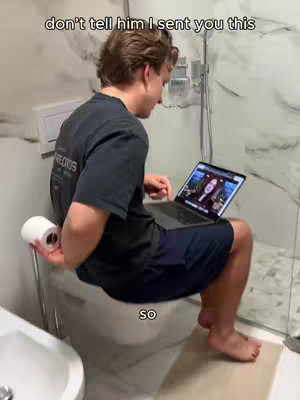 A man is sitting on a toilet seat holding a laptop, looking at the screen. A roll of toilet paper is in his other hand. The background shows a white-tiled bathroom with a shower visible.