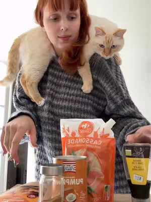 A woman with long red hair and a grey sweater stands indoors, with a cat perched on her shoulders. The cat is cream-colored with darker markings. In front of her on a table or kitchen counter are various food ingredients. The scene is brightly lit. 