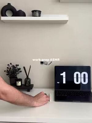 A clean, well-lit desk setup with a laptop displaying the time 1:00, various decorative items on a shelf, and a hand reaching towards the desk. The overall aesthetic is minimalist and focused, with a neutral color palette.