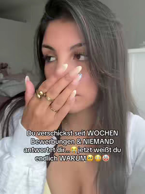 A young woman is holding her hand up to her mouth in a gesture that suggests surprise, or trying to cover her mouth. She has long, dark, wavy hair, and is wearing a white top and several rings. Text overlayed on the image reads: "Du verschickst seit WOCHEN Bewerbungen & NIEMAND antwortet dir...jetzt weißt du endlich WARUM." which translates to "You've been sending applications for WEEKS & NO ONE is answering you...now you FINALLY know WHY."