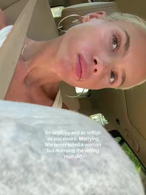 A selfie-style shot taken from inside a car, angled upwards. The primary focus is on a woman with fair skin and blonde hair. Her head is tilted. She is wearing jewelry and a white top. The text overlayed on the lower part of the image.