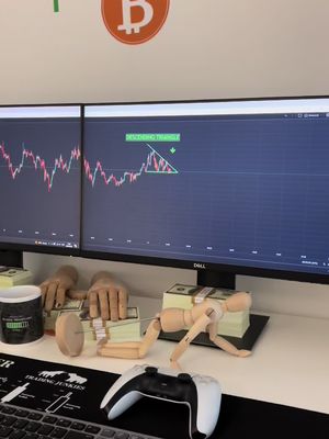 A desk setup featuring two computer monitors displaying stock charts, along with stacks of cash and an action figure. A gaming controller sits in the foreground.