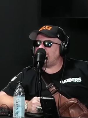 A man wearing sunglasses, a baseball cap with text, and headphones, holding a microphone in what appears to be a podcast setting. He is wearing a black shirt, and a water bottle and phone are visible in the foreground. The background is dark.