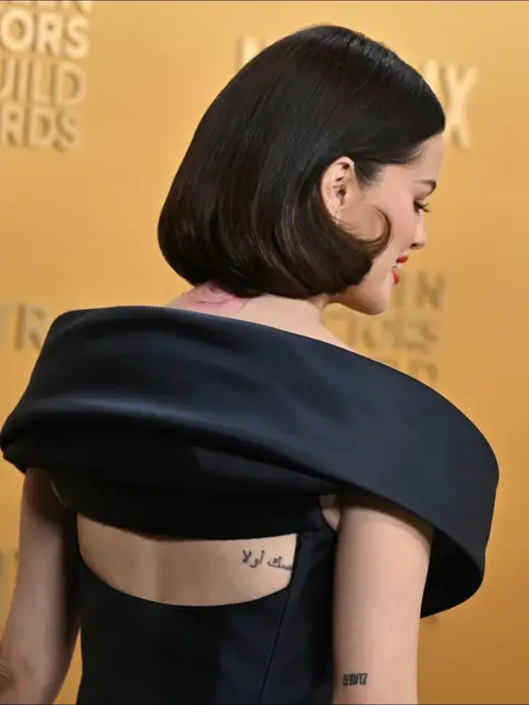 A side view of a woman with short, dark hair, wearing a black dress with a low back. The woman’s back displays two tattoos. The background is a solid tan color.