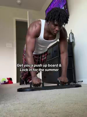 A man in a white tank top and plaid pants is in a room with a push-up board, getting ready to do pushups. Text overlay says, \"Get you a push up board & Lock in for the summer.\"