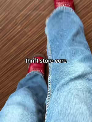 The image shows a low-angle shot of a person's legs wearing light blue jeans and red shoes. The jeans are faded with visible stitching along the seams. The red shoes have a studded detail. The background is a plain wood-like floor. The text 'thrift store core' is overlaid on the image along the leg.