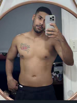 A shirtless man is taking a selfie in a mirror. He has a tattoo on his chest, a trimmed beard, and is holding a smartphone to capture the image.