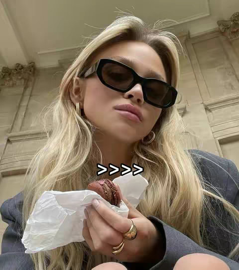A woman with long blonde hair is wearing black sunglasses and holding a dessert. The background is a blurred architectural detail.