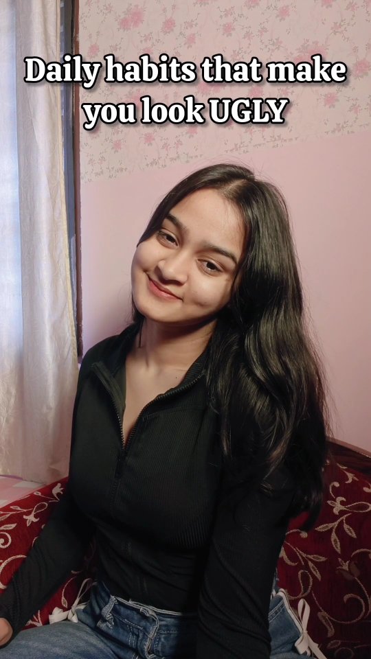 A young woman with long, dark hair is smiling at the camera, set against a soft pink wall with floral wallpaper. She wears a black zip-up top and jeans. The text overlay reads, 'Daily habits that make you look UGLY'.