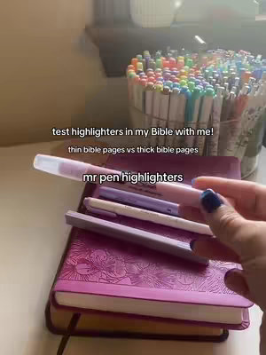 A video cover image shows a hand holding a pink highlighter over a Bible. Below, are a few other highlighters and a large container full of highlighters. There is visible text on the image: 'test highlighters in my Bible with me!' along with the text 'thin bible pages vs thick bible pages' and 'mr pen highlighters'. The setting appears to be indoors, possibly on a table next to a window. The overall tone is informative and practical.