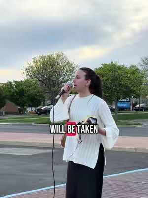 A woman with dark hair in a high ponytail is speaking into a microphone, holding a Bible. She is wearing a white sweater, and her black skirt and bag are visible, with greenery and cars in the background. Text overlay includes the phrase "WILL BE TAKEN".