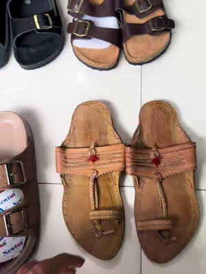 An overhead shot showcases several pairs of sandals resting on a light-colored, possibly tiled, surface. The sandals are in various styles and colors, including a pair of black slides, several with brown leather straps, and two pairs of open-toed leather sandals. The leather sandals have a more rustic, handmade appearance.