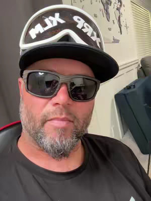 A man wearing sunglasses, a black hat, and a black shirt is the focus of a selfie-style shot. The backdrop includes a partially visible room with a neutral color scheme. The person has a beard. A pair of ski goggles are perched on the hat. The focus is sharp on the subject, creating a sense of closeness.