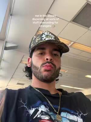 A selfie-style shot taken in what appears to be an airport. The focus is on a young man with a beard and a camouflage baseball cap. The background includes ceiling tiles and a window.