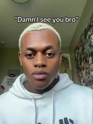 A medium shot shows a young person of color with short dyed blonde hair and a nose ring. The person wears a white hoodie. The background includes a wall with a collection of images and a circular ceiling light. The overlay shows text at the top that says "Damn I see you bro".