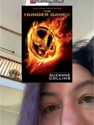 The cover image features a partial face shot, focusing on the eyes and lower face of a person with purple hair. The person is close to the camera with the book cover "The Hunger Games" book visible behind her. The background also shows a visible portion of a staircase and railing.