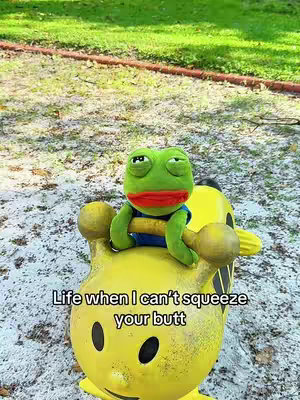 The image features a plush frog character sitting atop a yellow object resembling a Pokemon character. The frog is green with red lips, wearing a blue shirt and looking to the left. The frog is positioned amidst blurred green and brown foliage.