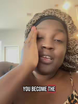 A medium close-up shot of a woman wearing a patterned headscarf and a leopard print top. The woman is looking slightly off-camera with her right hand raised to touch her forehead. Below the woman's face, the text overlay reads, "YOU BECOME THE". The background is softly lit with a hint of an interior setting visible.