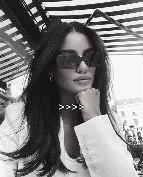 A black and white photo of a woman wearing sunglasses and a white blazer. The woman is looking downward with her hand by her chin and a white sign with arrows is over her chest, in front of the sunglasses.